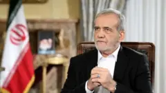 عکس مسعود پزشکیان رئیس جمهور ایران - با یک کورتی سیاه و پیراهن سفید در حالی که پرچم ایران در گوشه چپ تصویر دیده می‌شود