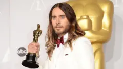 Jared Leto in 2014, wearing a white suit and satin red bow tie, poses for cameras holding up his Oscar award with one hand. He is against a white background and there is a large gold replica of the award behind him. 