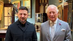 黒い襟付きの服を着たゼレンスキー氏と、グレーのスーツを着たチャールズ国王が、豪華な調度品や絵画が飾られた廊下に立っている写真