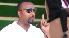 ቀዳማይ ምኒስተር ኣቢይ ኣሕመድ መደረ እናስምዐ። መነጽር ወድዩ ኢዱ ሓፍ ኣቢሉ ይርአ።