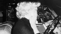 Imagem em preto e branco de Marilyn Monroe, sentada em um carro com as mãos cobrindo o rosto. Ao fundo, a mão de um homem está sobre o volante