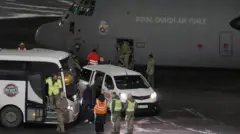 地面近くまで胴体を下げた灰色の機体に「王立デンマーク空軍」と白い文字で書かれた輸送機から、迷彩服姿の兵士たちが降りてきている。手前に、空港内の移動用車両が停まり、蛍光色のベストを着た人たちが、白い氷の見える駐機場に待機している