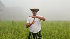 সবুজ ধানখেতের ভেতর কালো প্যান্ট, সাদা টিশার্ট ও ক্রিকেট আম্পায়ারদের মতো টুপি পরে দুই হাতে রিভিউ আবেদন দেখাচ্ছেন বিএনপির প্রার্থী