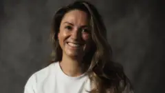 Imagem de uma mulher sorrindo. Ela tem cabelos claros e usa uma camisa branca