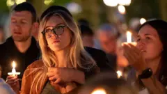 Seattle'da Charlie Kirk için düzenlenen anma nöbetinde bir kadın, arkadaşı kendisine arkadan sarılmış üzgün bakıyor. Fotoğrafta çok sayıda mum var.