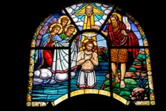 Vitral ilustrando o Batismo de Jesus. A figura central de Jesus está nas águas azuis do Jordão, enquanto João Batista, vestido com peles de animais, realiza o rito. Três anjos alados com vestes brancas aguardam à esquerda, e uma pomba, representando o Espírito Santo, desce de uma luz divina acima. A obra de arte em arco apresenta cores ricas e vibrantes e a tradicional escrita Ge'ez na base.