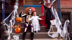 bir evin basamaklarında iki çocuğuyla oturan cadılar bayramı kıyafeti giymiş bir anne 