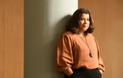 Luciana Chong posa encostada em uma parede marrom com semblante neutro e as mãos nos bolsos da calça preta. Veste uma blusa terracota e usa um colar longo