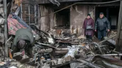 Чоловік риється у завалах зруйнованого після обстрілу будинку, позаду стоять чоловік та жінка в зимовому одязі