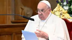 Pope Francis sitting in a velvet chair, reading from a sheet of paper. He is speaking into a microphone. The background is blurred.