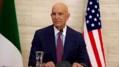 Kel, takım elbiseli Tom Barrack mikrofon başında. Arkasında Amerikan bayrağı var.