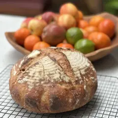 Pão de fermentação natural caseiro sobre uma mesa, ao lado de uma cesta de frutas