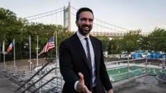 Zohran Mamdani estende a mão. Ele está de terno. Atrás dele, vê-se uma ponte, junto com bandeiras.