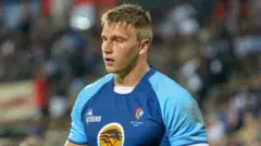 Rayno Nel, jovem adulto, visto do peito para cima, aparentando cansaço durante uma partida de rugby. Ele veste uniforme com o peito em azul royal, mangas em azul celeste e o logo do patrocinador em mostarda. Ao fundo, uma multidão desfocada.