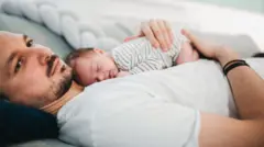 Un padre descansa en la cama con su bebé dormido sobre el pecho.