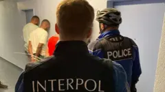 Two men wear interpol uniform stand wit three other men wey wey face wall