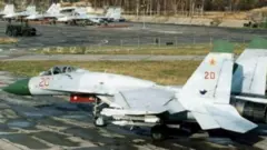 Винищувач Су-27 (Сухой), світло-сірого кольору з зеленим носом стоїть на бетонній стоянці аеродрому. На борту червоний номер "20" і червона зірка на хвості. Під крилом видно підвісні вузли для озброєння. На задньому плані – кілька інших літаків, припаркованих на бетонній площі.