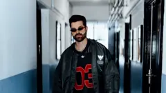 Alok, um homem de barba e cabelo escuro, posa em um corredor de paredes brancas e azul-acinzentadas, olhando diretamente para a câmera com expressão séria. Ele veste um casaco oversized preto sobre uma camisa esportiva preta com o número "03" em vermelho, além de óculos de sol estreitos e um colar prateado, compondo um visual de inspiração urbana e contemporânea.