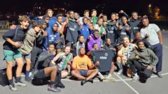 Environ 25 personnes se rassemblent pour une photo de groupe sur un terrain de football en béton.