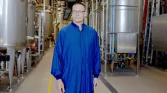 Um funcionário da fábrica da Novo Nordisk,m vaidebetpele clara e usando óculos, veste um macacão azul enquanto posam vaidebetfrente a tonéism vaidebetmetal dentro da unidade.