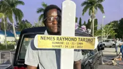 Umugabo ufashe umusaraba inyuma y'imodoka irimo umurambo wa Tarimo