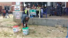 Aworan yii ṣafihan ọkunrin kan to bẹrẹ to si n sọ iwe idibo rẹ sinu apoti idibo, awọn eleto idibo naa wa lapa oke, wọn jokoo, wọn kawọ le tabili kan. Awọn agbofinro wa nibẹ pẹlu, bẹẹ si ni awọn to n fẹsẹ rin kọja tun wa ninu aworan idibo ijọba ibilẹ to waye nipinlẹ Rivers ni Satide, ọgbọnjọ oṣu Kẹjọ ọdun 2025.