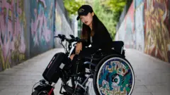 Uzun koyu renk saçlı, siyah şapkalı ve tekerlekleri grafitilerle süslenmiş bir tekerlekli sandalyede oturan bir kadın. Kadın, beton bir yolda, açık havada resmedilmiş ve her iki tarafındaki duvarlarda da grafitiler var.