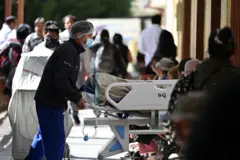 Un enfermero con tapabocas y malla de pelo lleva a un paciente en una camilla por un corredor de un hospital en el que hay mucha gente