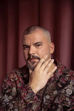 Retrato de um homem de cabeça raspada e barba curta, com expressão séria, olhando para a câmera. Ele apoia a mão no rosto, tocando a bochecha, e veste uma camisa estampada em tons de vermelho, dourado e preto. Ao fundo, há uma cortina vermelha que cria um contraste quente com a iluminação suave do retrato.