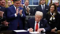 ダークスーツに赤いネクタイを着けたトランプ米大統領が、予算案に署名している。まわりには、ダークスーツに赤いネクタイ、めがねを着けたジョンソン下院議長（共和党）らが集まり、笑顔で拍手している