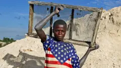 Un jeune garçon, en T-shirt aux couleurs du drapeau américain, se tient près d’une mine artisanale tenant un plateau de tamisage derrière ses épaules à Lumumbashi en RDC, décembre 2005.