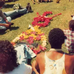 Pessoas sentadas em gramado em volta de túmulo; flores formam o desenho de uma guitarra