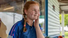 Uma jovem jogadora de críquete, com os cabelos ruivos trançados sobre o ombro, aplica protetor solar no rosto.