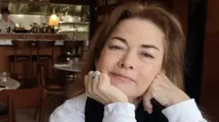 Sentada em um restaurante com iluminação baixa, Caroline olha para a câmera com expressão neutra. Vestindo uma blusa branca, apoia a cabeça na palma da mão direita. Ao fundo, é possível ver um chef na cozinha