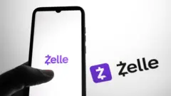 Uma mão segurando um celular com o logotipo do aplicativo Zelle na tela, com letras roxas sobre um fundo branco. Ao fundo, é possível ver o mesmo logotipo, nas cores preto e roxo