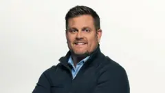 A foto mostra um homem, Brian Thompson, sorrindo, de braços cruzados, usando uma camisa azul-clara sob uma jaqueta escura, diante de um fundo branco.