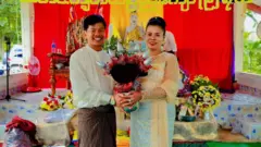 ကထိန်အလှူပွဲစာတန်းနောက်ခံနဲ့ ဘုရားဆင်းတုရှေ့မှာ ကထိန်သင်္ကန်းထည့်ထားတဲ့ ဖလားကို ကိုင်ပြီး ဓာတ်ပုံရိုက်ခံနေတဲ့ ဆရာမဒေါ်အေသီအောင်နဲ့ ခင်ပွန်း ဦးပြည့်ဖြိုးနိုင်