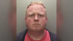 Robert Bush, com cabelo curto e castanho-claro, aparece em uma foto de ficha policial tirada de frente. Ele tem expressão neutra e veste uma jaqueta escura sobre uma camisa vermelha. O fundo é simples e cinza.