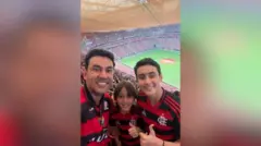 Thales Machado com os dois filhos, todos vestem camisas do Flamengo e sorriem olhando para a câmera, na torcida de um estádio de futebol lotado
