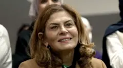 Close-up of Hind Kabawat, smiling. She has shoulder length brown hair and is wearing a smart, light brown coat.