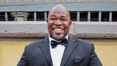 A man smiles broadly at the camera, wearing a black tuxedo and white shirt against a yellow and grey wall 