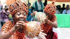 Two weomen wey dey smile wear beaded croen and calabash dey dance, dem dress like di Benin pipo from souther Nigeria.