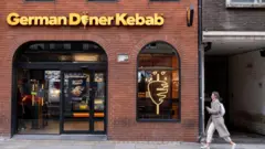 Fotoğrafta Alman Döner Kebap adıyla kurulan restoran zincirinin bir şubesi var. Bir kadın dönercinin önünden geçiyor.