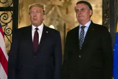 Donald Trump vistiendo una chaqueta oscura, camisa blanca y corbata roja está de pie junto a Jair Bolsonaro, quien viste una chaqueta oscura, una camisa azul con una corbata oscura con rayas claras.