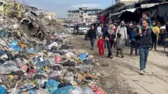 Palestinos desplazados caminan por un mercado junto a un vertedero de basura cubierto de bolsas de plástico. Un hombre pasa con un niño sobre los hombros, ambos mirando el vertedero. Detrás de ellos avanzan varios hombres, mujeres y niños. Al fondo se ven furgonetas blancas y edificios en ruinas.