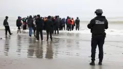 浮き具を身につけた人々が浅瀬に立っている。近くで警官が海の方を眺めている