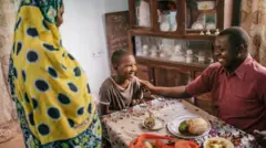 Man sidon wit pikin. Plate of food dey dia front and woman stand dey look dem