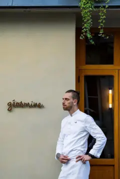 A foto mostra um homem jovem, de pele clara, vestindo uma doma branca, de perfil em uma parede onde está escrito Géosmine.