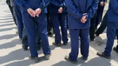 Спини чоловіків, що стоять у шерензі в темно-синій формі, тримаючи одну руку за спиною.