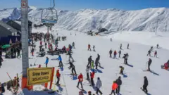 Skiers and snowboarders wait to join ski lifts at the Gudauri resort in Georgia. The ground is covered in snow and there are snow-capped mountains in the background